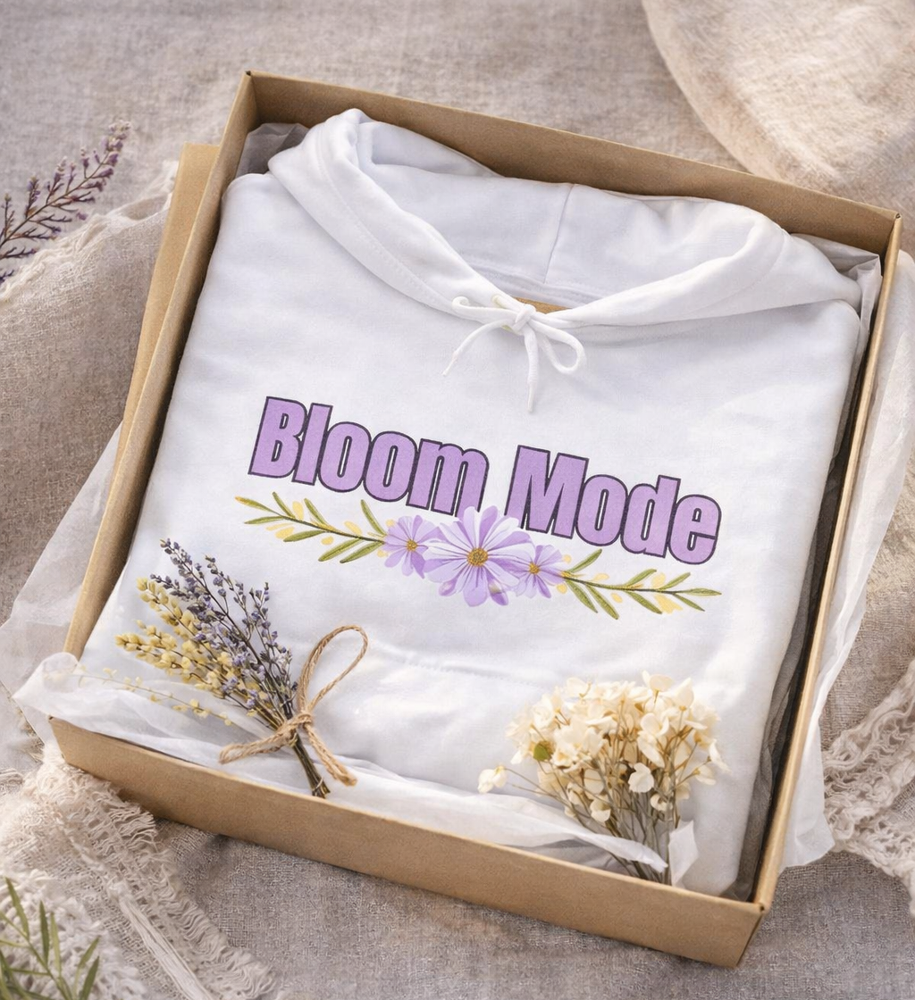 Weißer Unisex-Hoodie mit dem Schriftzug „Bloom Mode“ und einem floralen Blumenmotiv in Lilatönen auf der Vorderseite.