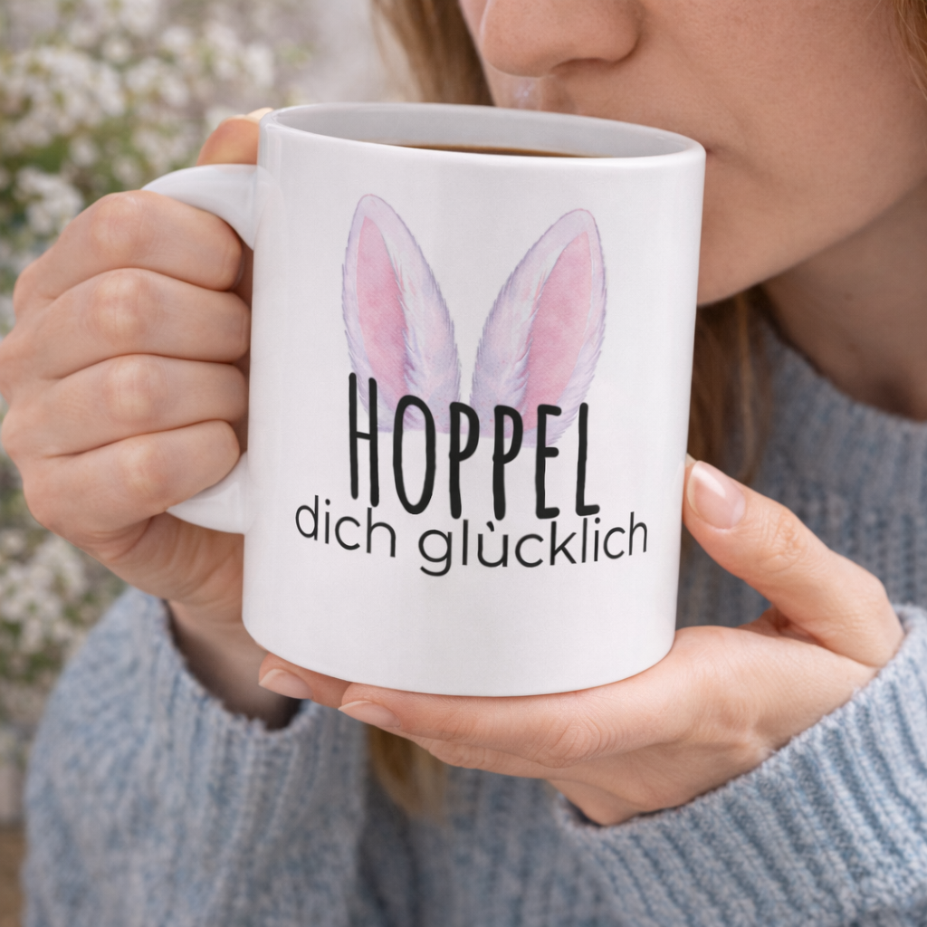Weiße Keramiktasse mit dem Spruch „Hoppel dich glücklich“, rosa Hasenohren sowie dem Schriftzug „Frohe Ostern“ und einem niedlichen Osterhasen-Design – 300-ml-Tasse.