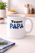 Weiße 300-ml-Keramiktasse mit dem Schriftzug „Team Papa“ in Blau und Schwarz sowie dekorativen Schnurrbart-Motiven – modernes Geschenk für Väter.